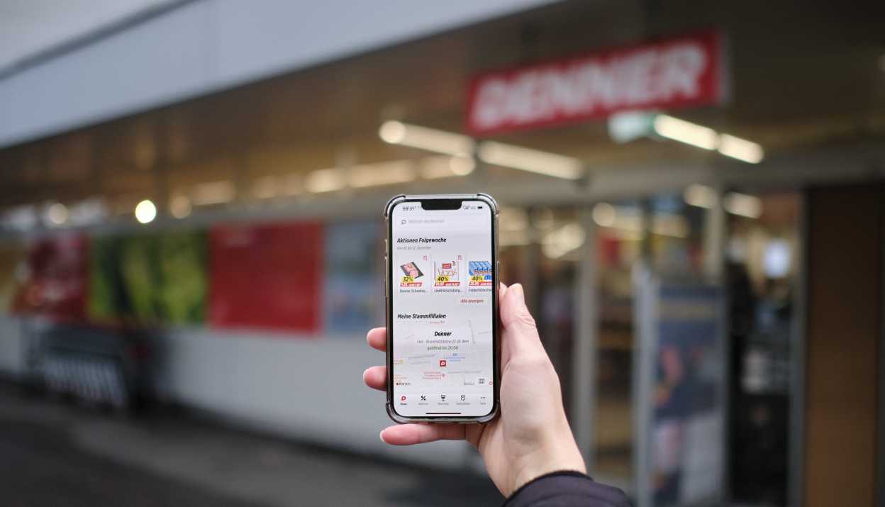 Cell phone with open Denner app in the hand of a person in front of Denner store