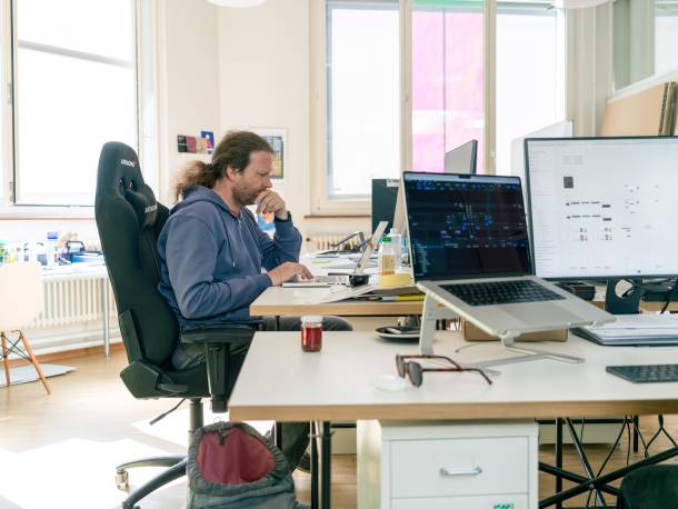 Olivier Oswald is working on his laptop. In the foreground are several monitors with open programmes. In the background is a large window.