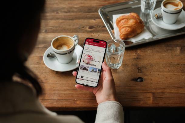SWIplus App geöffnet auf einem Smartphone in der Hand einer Person, die an einem Holztisch sitzt auf dem zwei Kaffees, zwei Gläser und ein Croissant stehen