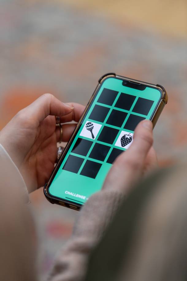 Nahaufnahme einer Person, die ein Smartphone in beiden Händen hält und eine App bedient. Auf dem türkisfarbenen Bildschirm ist ein Raster mit verdeckten Feldern sowie aufgedeckten Symbolen, darunter eine Erdbeere und eine Rassel, zu sehen.