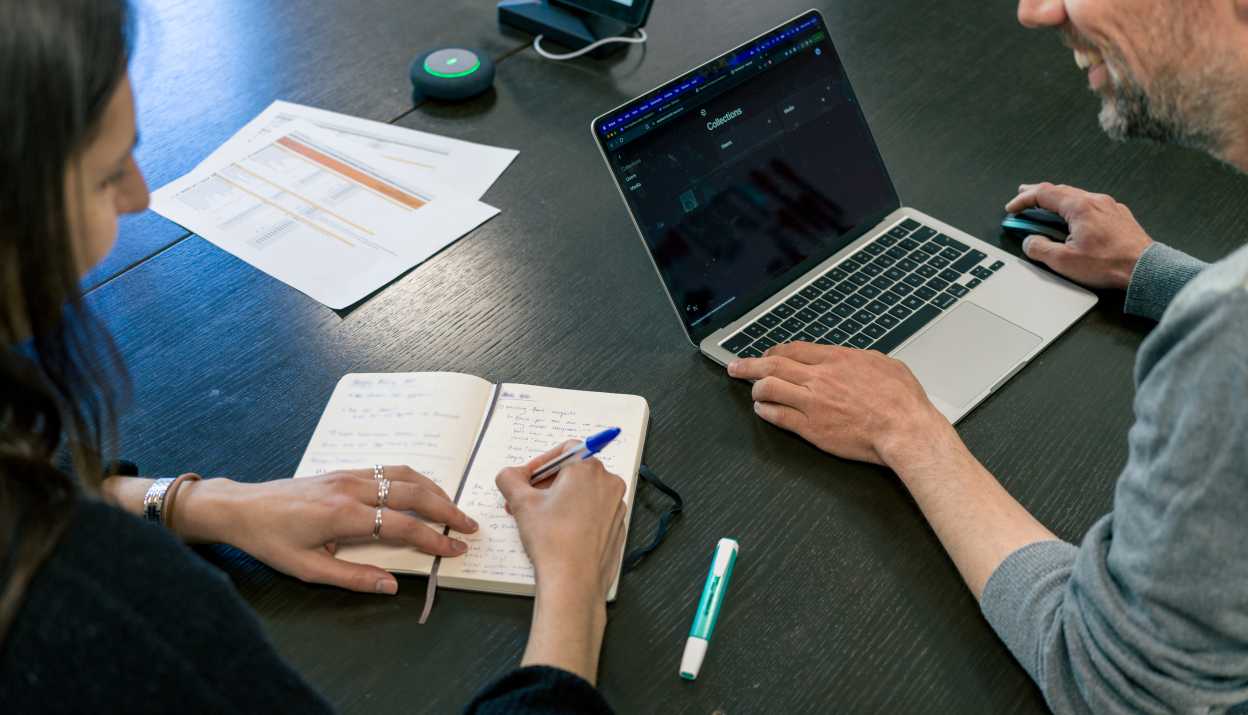 A woman and a man are sitting next to each other at the table, she is taking notes in a notebook, he has his laptop in front of him