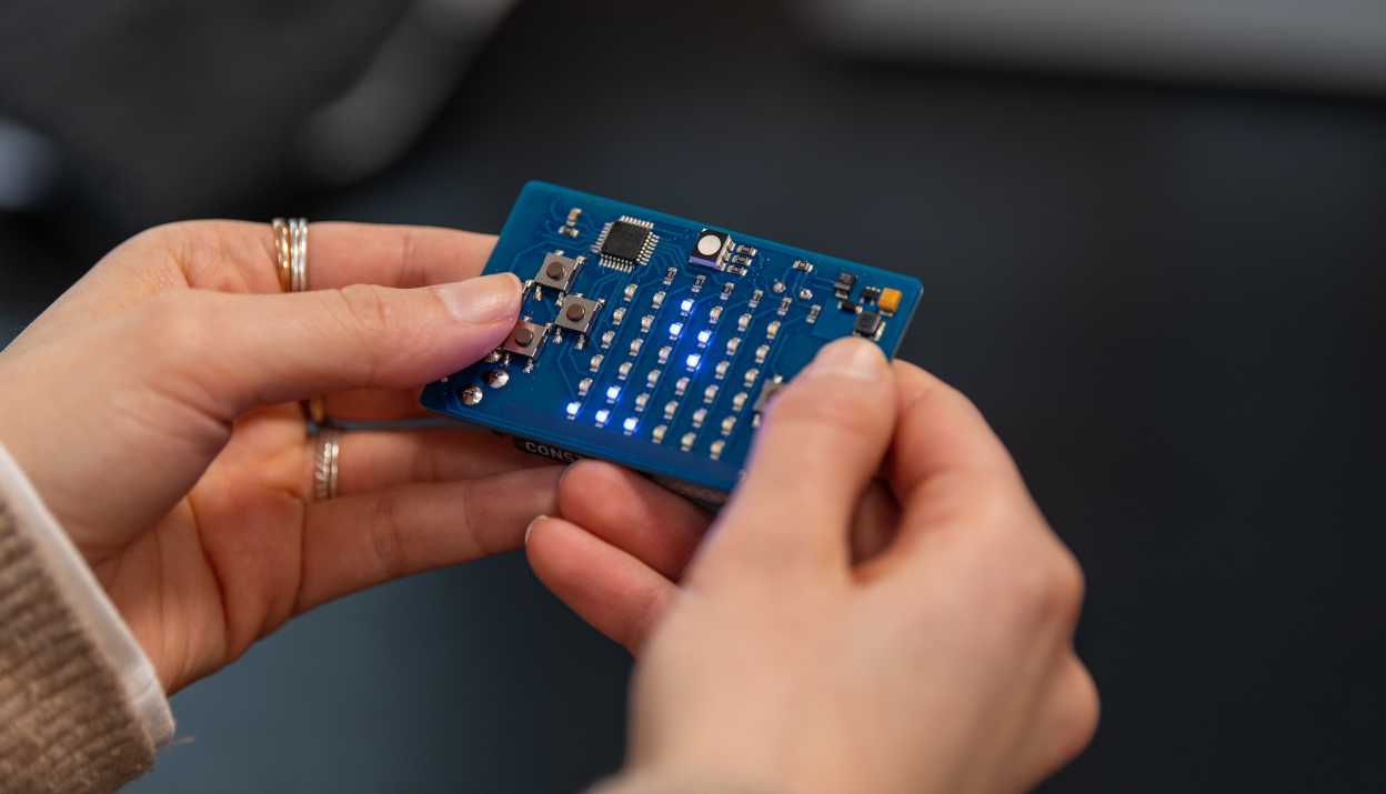 Close-up of two hands holding a small blue electronic circuit board with several LEDs lit up.