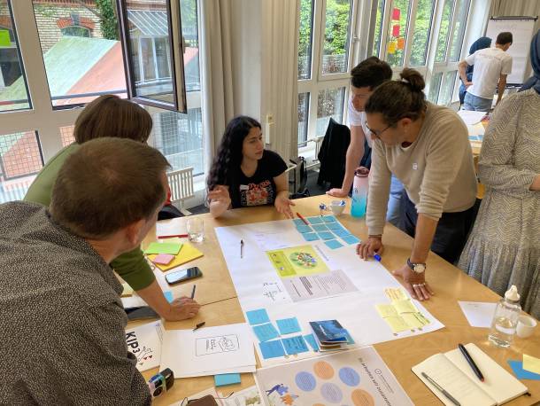 Personen stehen und sitzen rund um einen Tisch, sammeln auf Post-its Ideen und diskutieren